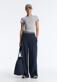 Kvinde i grå tætsiddende t-shirt, marineblå bukser med brede ben og stribet taljebånd, olivengrøn baseballkasket, holder en marineblå tote taske og har sneakers på.