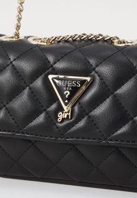 Sac en bandoulière matelassé noir avec une chaîne dorée. Il présente une forme structurée et un logo triangulaire proéminent avec les accents "GUESS" et "girl".