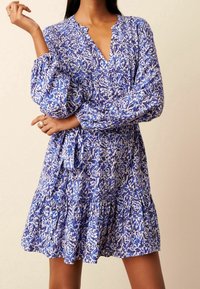 Sininen ja valkoinen kuviollinen mekko, jossa on pitkät hihat, V-pääntie ja kerroksellinen helma, valmistettu kevyestä kankaasta.