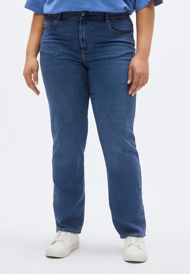 Mørkeblå denimjeans med rett snitt, fem lommer og diskrete sømdetaljer; kombinert med hvite joggesko.