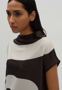 Top a righe bianche e nere con scollo alto, texture di raso liscio e maniche corte. Presenta blocchi di colore audaci e una vestibilità rilassata.