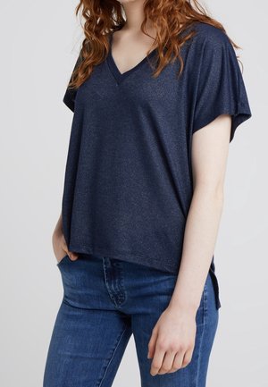 Femme portant un haut bleu marine à col en V et manches courtes, ainsi qu'un jean bleu, mains dans les poches, sur un fond uni.