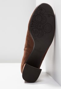 Semelle de chaussure en daim marron avec des motifs circulaires embossés et un talon bloc reposant contre des surfaces blanches.