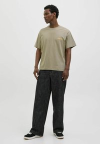 Oliven grøn bomulds T-shirt med korte ærmer og gul logo, parret med løse, mørkegrå denimbukser og sorte sneakers med hvide snørebånd.