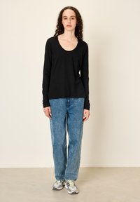 Kiharaliste juustega naine, kes kannab musta pika varrukaga särki, siniseid teksaseid ja halli spordijalatsit, seisab ühevärvilise beeži tausta ees.