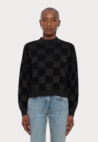 Pull cropped noir et or à carreaux avec un col montant et des manches longues, présentant un motif tricoté texturé. Porté avec un jean bleu clair.