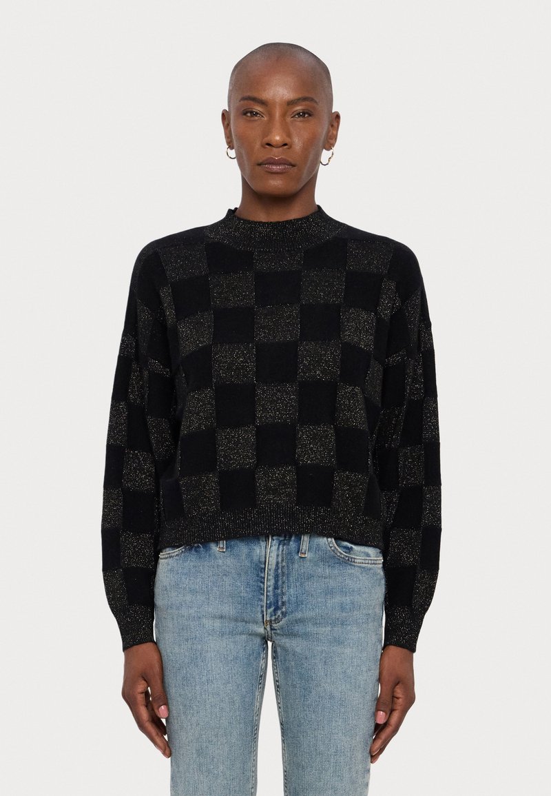 Pull cropped noir et or à carreaux avec un col montant et des manches longues, présentant un motif tricoté texturé. Porté avec un jean bleu clair.