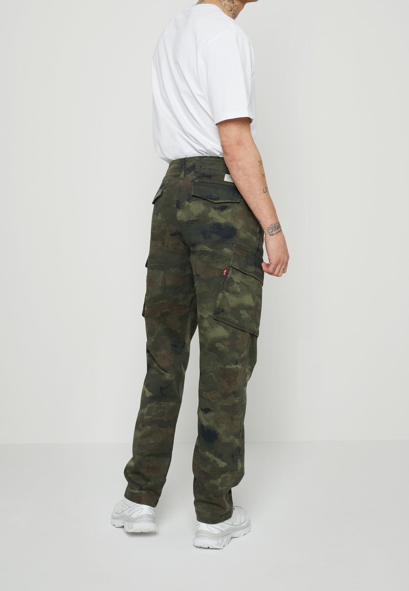 Personne portant un t-shirt blanc et un pantalon cargo camouflage vert avec des baskets blanches, debout de dos sur un fond uni.