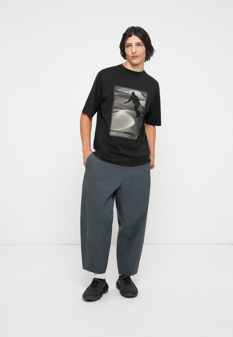 T-shirt nera a maniche corte con stampa grafica, abbinata a pantaloni larghi grigi. Il modello indossa scarpe da ginnastica nere, in piedi su uno sfondo neutro.