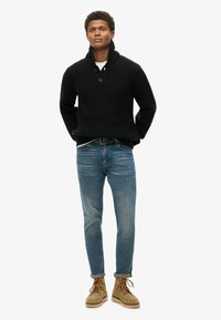 Schwarzer Strickpullover mit Schalkragen und zwei Knöpfen, kombiniert mit blauen Jeans und hellbraunen Schnürstiefeln.