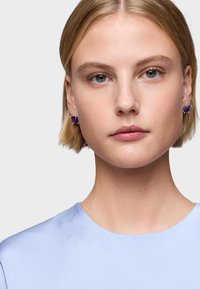 Orecchini in gemma viola montati in argento, con forma rettangolare. Il modello indossa un top satinato azzurro chiaro con scollo rotondo. Capelli corti.