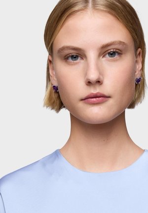 Pendientes de gema púrpura engastados en plata, con forma rectangular. La modelo lleva una blusa de satén azul claro con cuello redondo. Pelo corto.