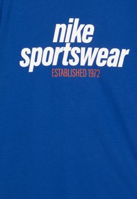 Zila kokvilnas T-kreklu ar izteiksmīgu baltu "nike sportswear" tekstu un oranžu "IZVEIDOTS 1972" detaļu. Gluda tekstūra, ikdienišķs dizains.