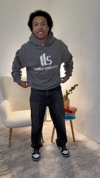 Sweatshirt cinzento com capuz e logótipo branco, calças pretas e ténis pretos e brancos. Em pé num interior bem iluminado com fundo de cor clara.