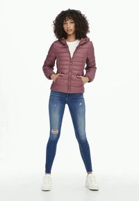Mulher com cabelo encaracolado vestindo uma jaqueta acolchoada em tom mauve, jeans skinny rasgados e ténis brancos, de pé com as mãos nos bolsos.