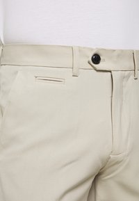 Beige byxor med en slät textur, som har en svart knapp, dragkedja och en diskret insticksficka fram.