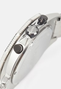 Reloj de acero inoxidable plateado con un bisel texturizado, que cuenta con dos coronas: una con un detalle negro y marca, y una parte trasera de caja lisa.