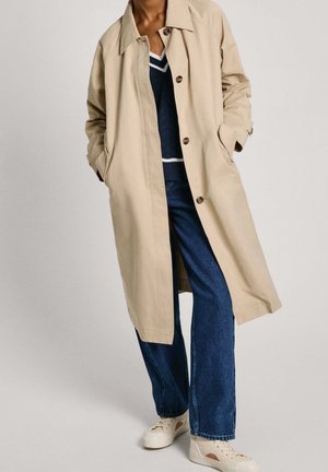 Beige trench coat, jossa on rento leikkaus, varustettu lovikolarilla, etunapeilla ja sivutaskuilla, käytetään tummansinisen villaneuleen ja sinisten farkkujen päällä.