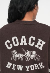 Sweater de malha castanho com texto branco "COACH NEW YORK" e uma imagem de uma carruagem puxada por horse. Apresenta um decote canelado e costuras detalhadas.