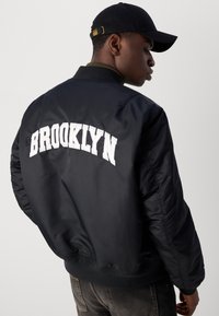 Crna bomber jakna s bijelim izvezenim natpisom "BROOKLYN" na leđima. Ima rebrasti ovratnik i manšete, te glatku, sjajnu teksturu.