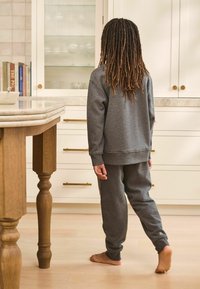 Una felpa grigia con cappuccio e pantaloni felpati sono indossati, con un taglio rilassato e polsini a coste. La persona è a piedi nudi, vista di spalle in una cucina.