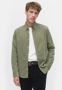 Oliivivihreä napillinen paita, joka on pehmeästä puuvillasekoitteesta, jossa on terävä kaulus, yksi rintatasku ja valkoiset napit, yhdistetty valkoisen t-paidan kanssa.