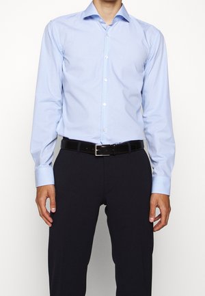 Man wearing light blue checkered button-up shirt tucked into black pants with a black leather belt, standing against a white background.