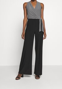 Svart overall med ärmlös design, med ett mönstrat överdel, omslagsfront och knytning i midjan, tillverkad av mjukt tyg.