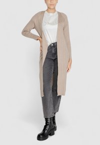 Cardigan beige a costine con maniche lunghe e apertura frontale, abbinato a una t-shirt grigia chiara e jeans neri a vita alta, completato da stivali neri chunk.