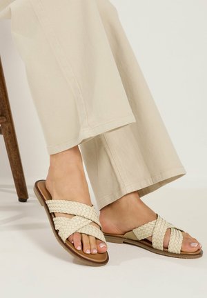 Voeten met beige gevlochten lederen instapsandalen met brede bandjes, gecombineerd met lichtgekleurde wijde broekspijpen.