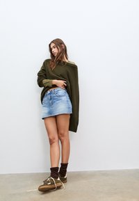 Pull oversize tricot vert olive, mini-jupe en jean bleu, chaussettes marron et chaussures vertes avec accents blancs sur un sol lisse.