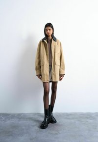 Femme portant une veste beige surdimensionnée avec un col noir, un pull marron, une jupe à motifs, des collants noirs transparents et des bottes noires brillantes, debout à l'intérieur.