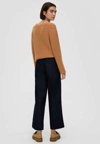 Camelowy sweter z dzianiny z długimi rękawami, zestawiony z ciemnoniebieskimi spodniami o szerokich nogawkach. Stylizacja charakteryzuje się luźnym krojem i podwiniętymi mankietami.