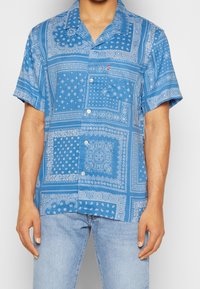 Chemise à manches courtes en tissu bleu avec un motif cachemire blanc, fermeture boutonnée à l'avant, et coupe décontractée. Idéale pour une tenue décontractée.
