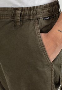 Close-up of hand in front pocket of olive green elastic waist pants with belt loop and small black label.