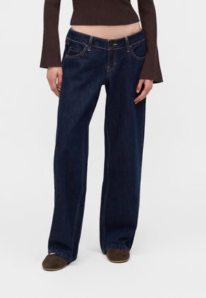 Personne portant un jean bleu foncé à jambes larges, un haut court marron côtelé à manches longues et des chaussures marron sur un fond uni.
