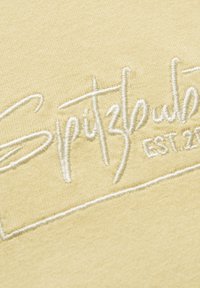 Ein cremefarbenes Gewebe zeigt ein in Weiß gesticktes Logo, das "Spitzbub" mit "EST. 20" darunter liest, vor einem strukturierten Hintergrund.