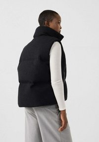 Persona che indossa un gilet imbottito nero sopra una maglia bianca a maniche lunghe a coste e pantaloni grigio chiaro, in piedi di profilo contro uno sfondo uniforme.