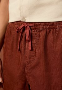 Pantalon en velours côtelé rouge profond avec une texture rayée, disposant d'une taille avec cordon de serrage et de poches latérales. Le tissu semble doux et confortable.