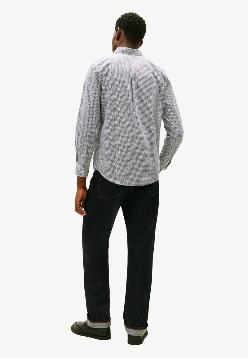 Homme debout, de dos, portant une chemise à manches longues à motifs, un jean foncé avec des revers et des chaussures noires sur fond blanc.
