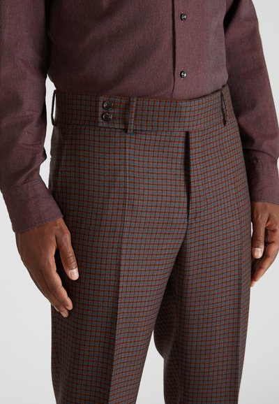 Man in bruin en blauw geblokte broek met een dubbele knoopband en een bordeauxrode longsleeve shirt, met handen langs de zij.