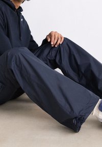 Personne portant un pantalon large bleu marine et un sweat à capuche assorti, assise par terre, les mains reposant sur le genou, chaussettes blanches et baskets visibles.
