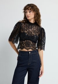 Sort blondecrop top med korte puffede ærmer, intrikat blomsterdesign, scallopkant, parret med mørk denim højtaljede bukser.