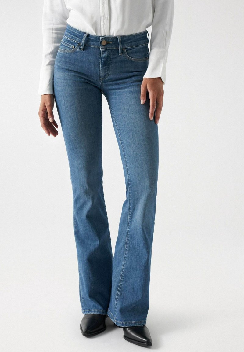 Femme portant un jean bootcut bleu, une chemise blanche à manches longues et des chaussures noires à bout pointu, debout devant un fond blanc uni.