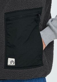 Fleece-Weste in Dunkelgrau mit einer schwarzen Reißverschlusstasche, versehen mit einem Logo-Label an der Unterseite. Strukturiertes Außenmaterial, glattes Taschenmaterial.