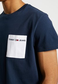 Námořnická modrá bavlněná trička s bílou náprsní kapsou a textem "TOMMY JEANS". Krátké rukávy a kulatý výstřih. Měkká textura.