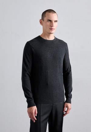 Mann mit kurzen Haaren, der einen dunkelgrauen Langarm-Pullover und dunkle Hosen trägt, steht vor einer schlichten hellgrauen Wand.