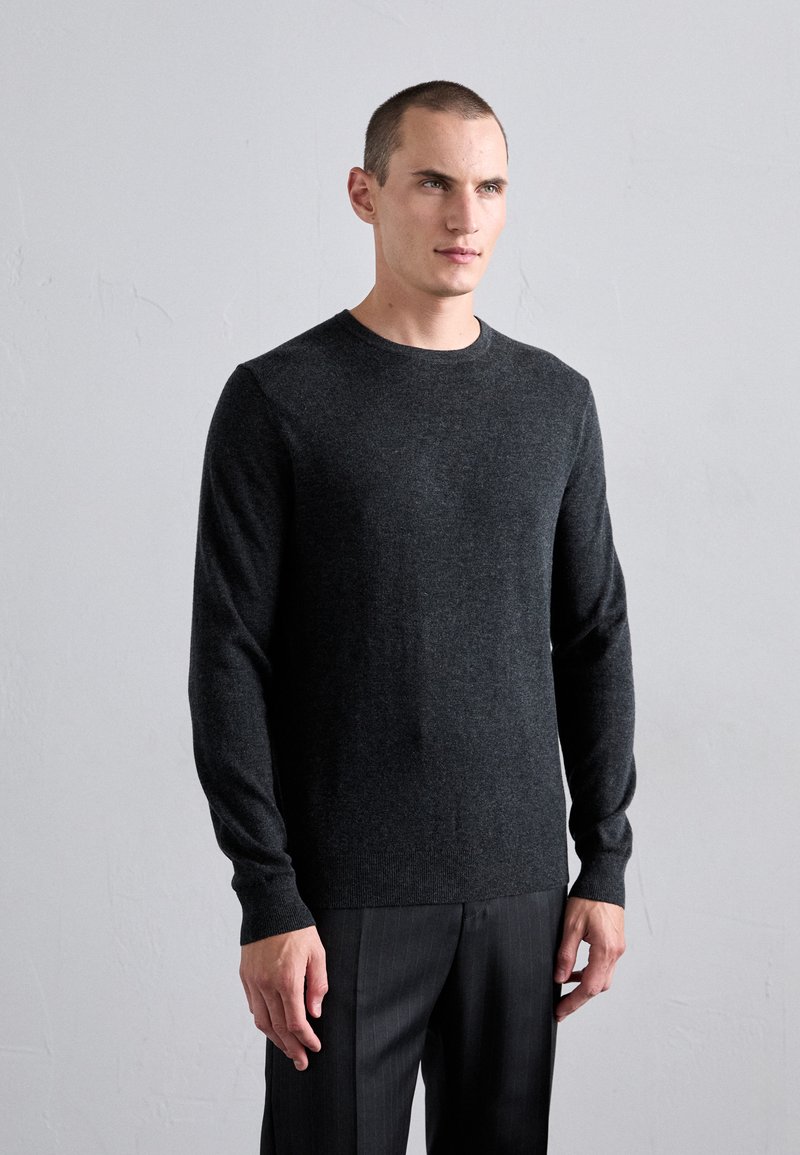 Man with short hair wearing a dark gray long-sleeve crewneck sweater and dark pants, standing against a plain light gray wall.