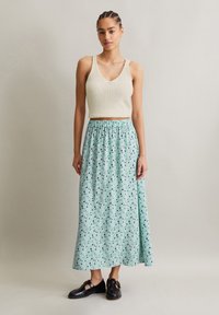 Débardeur en tricot crème avec un col en V, associé à une longue jupe verte menthe ornée d'un motif floral noir et vert foncé, complétée par des chaussures noires.