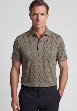 Man draagt een olijfgroen poloshirt met kleine donkere stippen en een zwarte broek, staand tegen een effen lichte achtergrond.